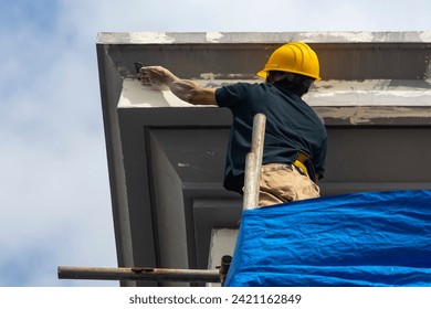 A skilled construction worker in a hard hat standing on top of scaffolding sanding the corner of the eaves and fascia of a 3 story house. Exterior renovation of a building facade.