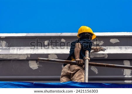 A skilled construction worker in a hard hat standing on top of scaffolding sanding the eaves and fascia of a 3 story house, in preparation for painting. Exterior renovation of a building facade.