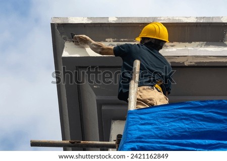 A skilled construction worker in a hard hat standing on top of scaffolding sanding the corner of the eaves and fascia of a 3 story house. Exterior renovation of a building facade.