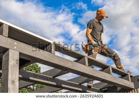 A skilled construction worker is standing on a wooden framework, carefully placing roof panels. The sunny day enhances the outdoor environment, showcasing the clouds.