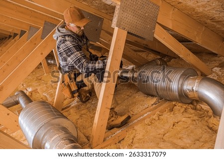 A skilled contractor is doing HVAC work in an attic, surrounded by insulation and ducting, using tools and focused on the task.