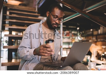 Skilled dark skinned programmer with coffee to go analyzing data code while working remotely on digital netbook, millennial IT professional in spectacles reading received email message on laptop