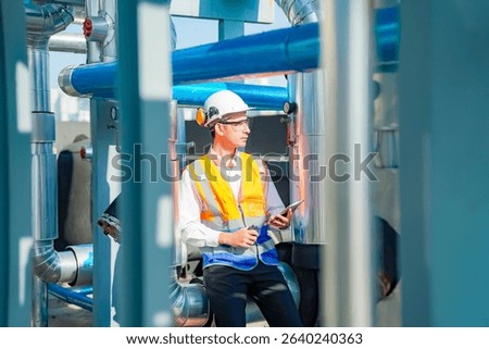 A skilled engineer is checking safety protocols while seated between large pipes in an industrial environment. He wears safety gear, including a hard hat and reflective vest, ensuring compliance.
