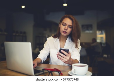 Skilled female administrative manager checking email box waiting for booking confirmation via smartphone connected to free wireless internet on coffee shop while planning business trip using laptop 