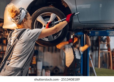 Skilled female auto mechanic standing near lifted car at car repair shop with tool in hands and changing tyre. Professional female technician standing at auto mechanic workshop and replacing tyre. - Powered by Shutterstock