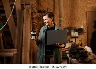 A skilled female carpenter holds a laptop and coffee cup, ready for a woodworking project in her workshop. - Powered by Shutterstock