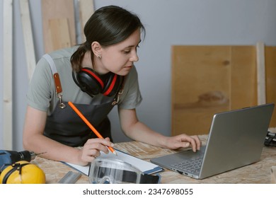 Skilled female carpenter making notes while using laptop at working table with carpentry tools. Young woman wearing apron working in craft workshop, learning online or checking order of clients.