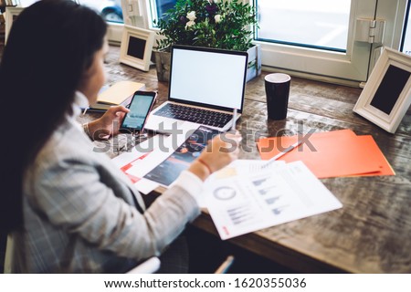 Skilled female student learning financial literacy from analytic documents for analyzing information and research results on modern smartphone gadget or blank laptop computer with copy space area