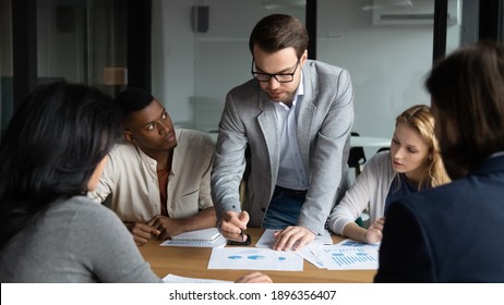 Skilled Focused Young Businessman Boss Leader In Eyeglasses Analyzing Sales Data Statistics Or Explaining Marketing Strategy To Motivated Diverse Multiracial Colleagues At Brainstorming Meeting.