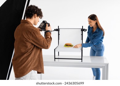 Skilled food photographer in brown jacket intensely focuses his camera on perfectly styled burger, with an assistant adjusting the presentation on white tabletop - Powered by Shutterstock