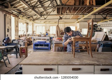 Skilled Furniture Maker Sanding A Wooden Chair On A Workbench While Working Alone In His Large Woodworking Shop