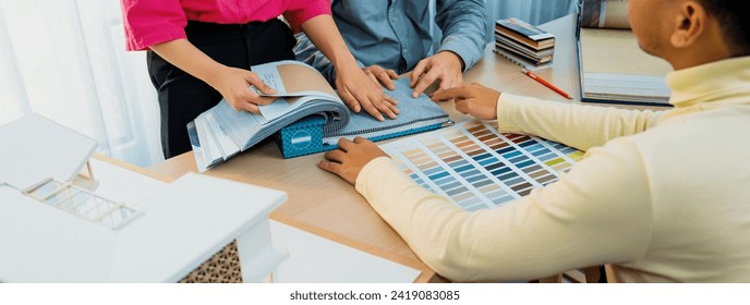 Skilled interior design team carefully selecting curtain materials while coworker selecting the color of curtain with house model placed on meeting table. Creative design concept. Variegated.