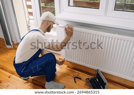 A skilled maintenance worker sitting close to a new radiator in a living room, ensuring its working efficiently to provide warmth during the cold winter months. Tools are nearby for seasonal service.