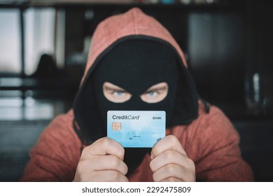 Skilled masked criminal using a stolen credit card to buy things online. A thief in a black mask holds a stolen bank card in his hands.