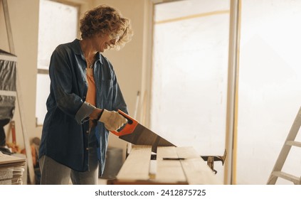 Skilled mature woman uses a crosscut saw to cut a wooden plank for baseboards. She is renovating the interior of a house, demonstrating her expertise in carpentry and home improvement. - Powered by Shutterstock