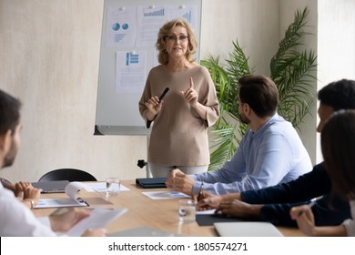 Skilled Middle Aged Older Female Leader Ceo Giving Educational Lecture Workshop Seminar Standing Near Whiteboard To Concentrated Young International Group Of Diverse Colleagues In Modern Boardroom.