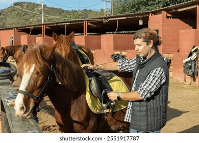 Skilled middle-aged male stable keeper checking saddle on brown horse for riding in yard of stables - Powered by Shutterstock