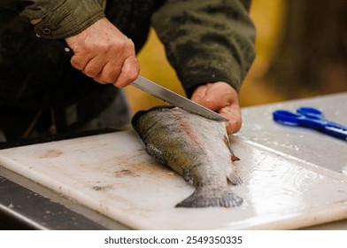 A skilled person fillets a fresh fish on a white cutting board outdoors, surrounded by vibrant fall colors. - Powered by Shutterstock