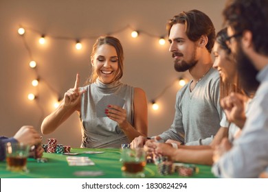 A Skilled Player. Smiling Young Woman Holds Up Finger As She Sees She Has Lucky Hand While Playing Game Of Poker With Friends, Sitting At Table With Drinks At Casino Themed Party At Home Or In Club