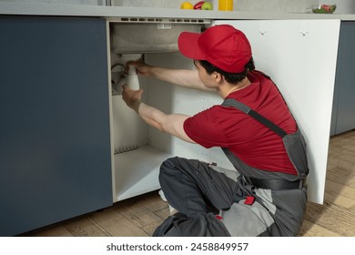 A skilled plumber in a red cap and uniform is diligently working to repair a kitchen sink pipe. Kneeling on the floor, the technician uses tools to fix the plumbing issue, ensuring the sink will - Powered by Shutterstock