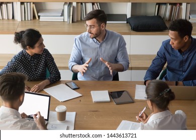 Skilled serious male manager talk to diverse business people at group meeting sit at conference table, multi ethnic team interns workers listen to mentor boss explain work plan in office, top view
