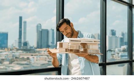 Skilled smiling caucasian engineer turns house model left and right to check mistake point. Professional manager inspect at architectural model while standing near window. Design concept. Tracery - Powered by Shutterstock