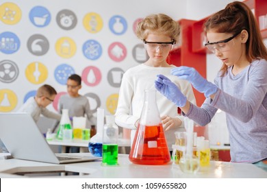 Skilled students. Pleasant schoolgirls in safety goggles conducting chemical experiment and thinking about the next step in it while their classmates having similar discussion in the background - Powered by Shutterstock