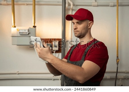 A skilled technician, wearing a red cap and overalls, is inspecting a heating system in the basement of a residential home. He appears focused as he uses a tablet to assess the systems performance