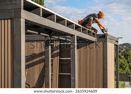 A skilled worker is constructing a sleek outdoor shed, carefully attaching wooden panels while surrounded by lush greenery and a clear sky.