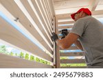 A skilled worker is focused on installing wooden slats in a backyard gazebo. The sun shines brightly, illuminating the workspace as the craftsman uses a power tool to secure the slats in place