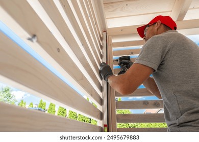 A skilled worker is focused on installing wooden slats in a backyard gazebo. The sun shines brightly, illuminating the workspace as the craftsman uses a power tool to secure the slats in place - Powered by Shutterstock