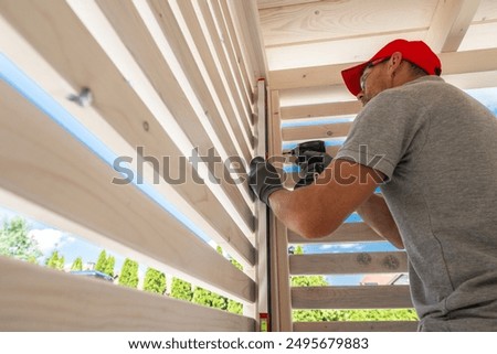 A skilled worker is focused on installing wooden slats in a backyard gazebo. The sun shines brightly, illuminating the workspace as the craftsman uses a power tool to secure the slats in place