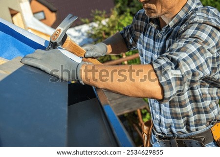 A skilled worker is installing roofing materials on a home’s upper section, using a hammer while wearing gloves. The sun shines brightly above, highlighting his focused effort.