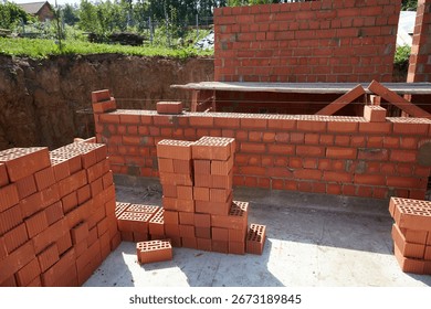 Skilled workers are constructing a brick wall with red bricks at a construction site. The structure is taking shape under clear skies, highlighting the building process. 