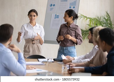 Skilled Young Arab Female Presenter Standing Near Flipchart With Happy Indian Colleague, Presenting New Marketing Strategy To Focused Mixed Race Business People Investors At Briefing Meeting In Office