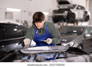 Skilled young auto body technician in garage, wearing blue overalls and green hoodie, applying filler to damaged car bumper, preparing for painting.. - Powered by Shutterstock