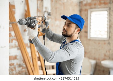 Skilled young builder in blue overalls working with pneumatic chisel hammer at indoor construction site..