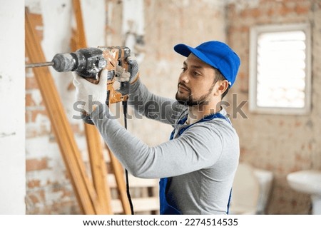 Skilled young builder in blue overalls working with pneumatic chisel hammer at indoor construction site..