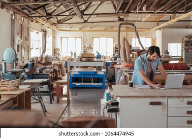 Skilled young craftsman standing at a workbench in his large carpentry studio full of woodworking equipment using a laptop - Powered by Shutterstock