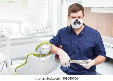 Skilled young dentist is sitting and holding medical tools. He is looking forward through special eyewear seriously. Copy space in left side - Powered by Shutterstock