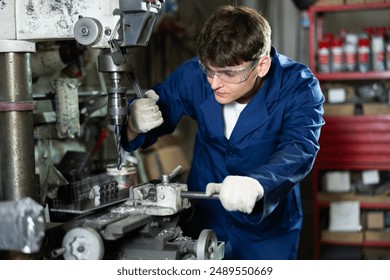 Skilled young factory worker, in blue work coat, protective glasses and gloves, working on drilling machine in processing workshop, performing holes in metal part - Powered by Shutterstock