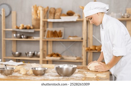 Skilled young female baker in white chef jacket and hat kneading dough on flour-dusted wooden table in warm inviting setting of small artisan bakery, showcasing traditional bread making skills - Powered by Shutterstock