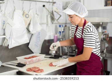 Skilled young female butcher in striped shirt and apron, using burger press to shape ground veal patties in processing room of local shop - Powered by Shutterstock