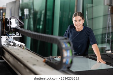 Skilled young female glass processing workshop worker in blue uniform working on CNC glass cutting machine, focusing on handling details - Powered by Shutterstock