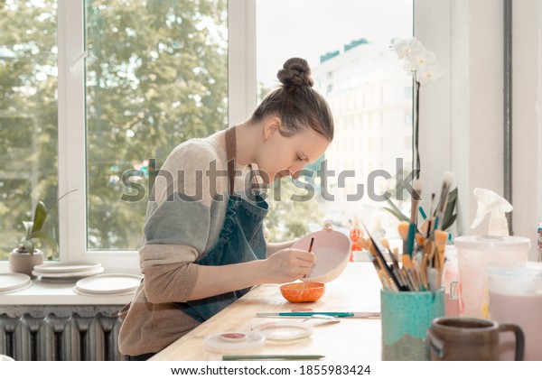 Skilled young woman in apron sitting at table\
and drawing on ceramic bowl in pottery workshop. Earn extra money,\
side hustle, turning hobbies into\
job.