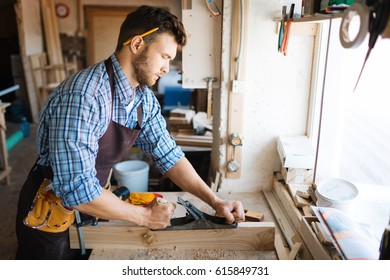 Skillful bearded woodworker in checked shirt smoothing plank with jointer plane, shavings scattered on table - Shutterstock ID 615849731