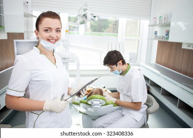 Skillful young dentist is examining teeth of child with concentration. The man is sitting near a boy with mask and eyewear. His assistant is standing and holding documents. Woman is smiling - Powered by Shutterstock