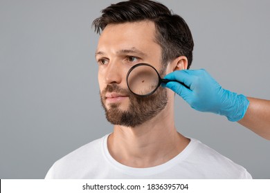 Skin Cancer Concept. Doctor Hand Checking Face Skin Of Middle-aged Man With Dermatoscope, grey studio background, copy space. Dermatologist making check up for bearded man, scanning mole on his face