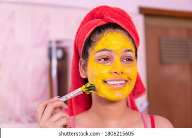 Skin care - Beautiful girl applying Gram flour turmeric yellow face mask on face through brush. She is wearing red towel on head.