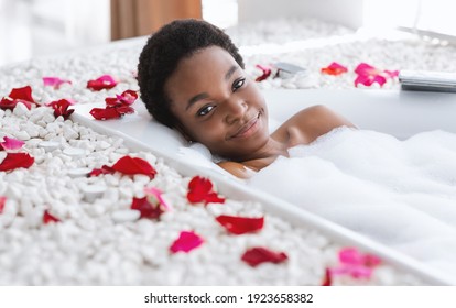 Skin care, clean body, spa treatments at home during covid. Calm smiling millennial african american lady in bathtub with foam and petals, enjoys water in bathroom interior in daylight, copy space